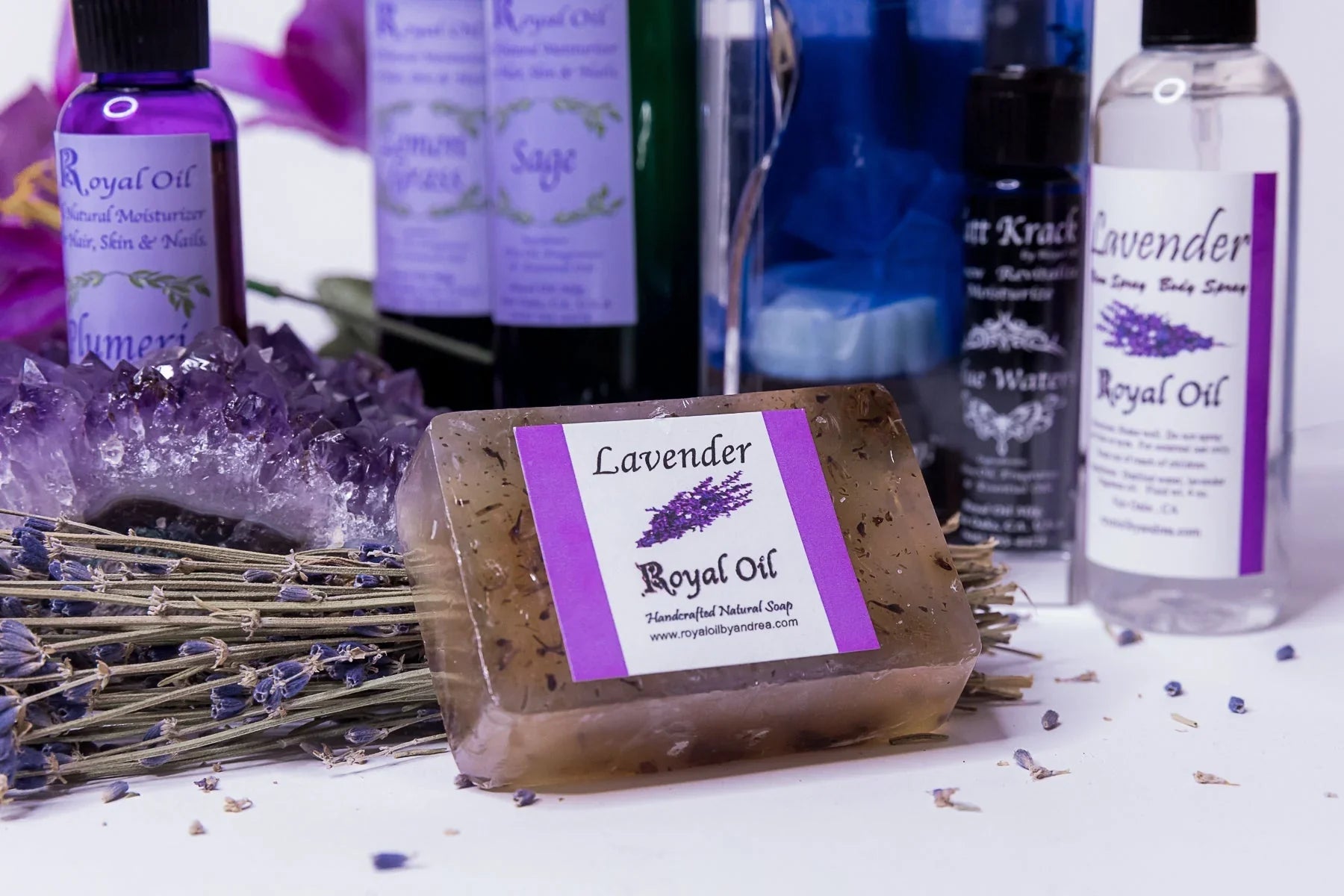A bar of Royal Oil Lavender handcrafted soap with botanicals sits on a white surface by a lavender bundle and amethyst. Royal Oil bottles of Plumeria, Sage, and Lavender spray are in the background