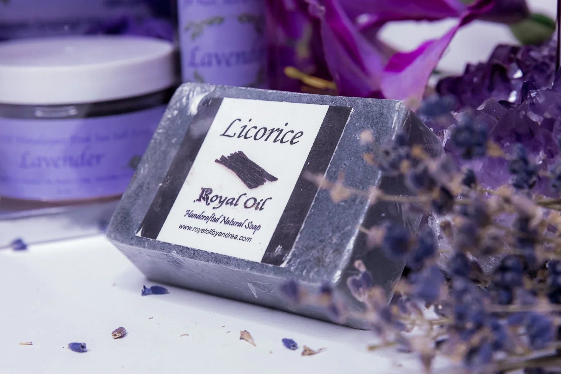Dark bar of Royal Oil Licorice handcrafted soap wrapped in plastic. It sits on a white surface scattered with lavender buds, surrounded by amethyst crystals, dried lavender, and purple skincare jars in a soft-focus background.